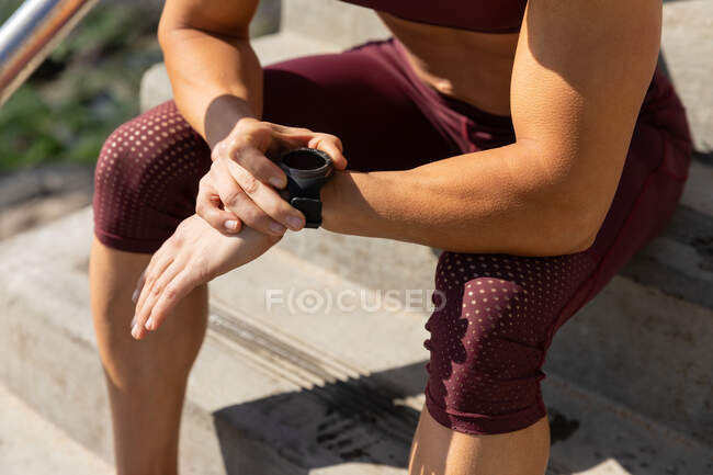 Seitenansicht einer sportlichen Frau, die an einem sonnigen Tag auf einer Strandpromenade trainiert, auf der Treppe sitzt und auf ihre Smartwatch blickt. — Stockfoto