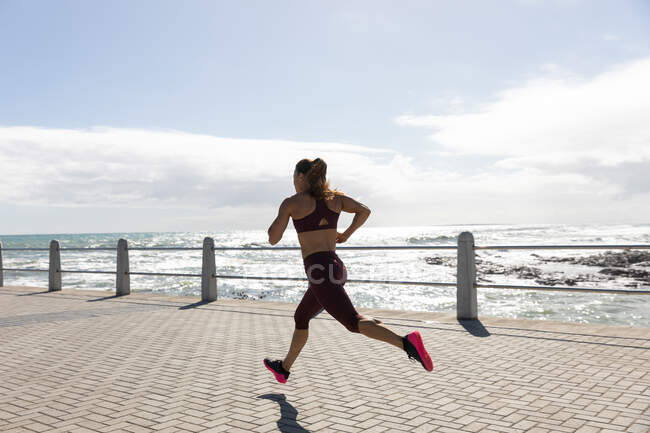 Vista lateral de uma mulher caucasiana desportiva com longos cabelos escuros se exercitando em um passeio à beira-mar em um dia ensolarado com céu azul, correndo . — Fotografia de Stock
