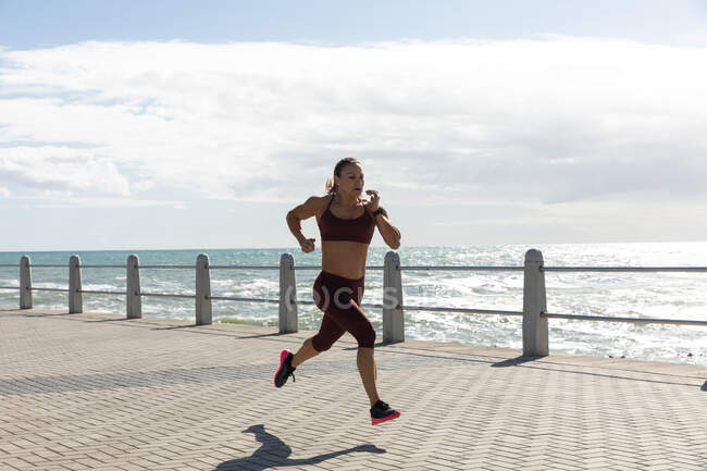 Vista lateral de uma mulher caucasiana desportiva com longos cabelos escuros se exercitando em um passeio à beira-mar em um dia ensolarado com céu azul, correndo . — Fotografia de Stock