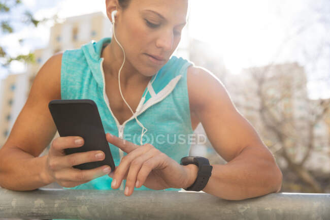 Front view of a sporty Caucasian woman with long dark hair exercising in the urban area on a sunny day, using her smartphone and checking her smartwatch with her headphones on. — Stock Photo