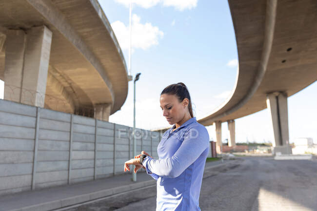 Vista lateral de una mujer atlética caucásica con el pelo largo y oscuro haciendo ejercicio en el área urbana en un día soleado, revisando su reloj inteligente. - foto de stock
