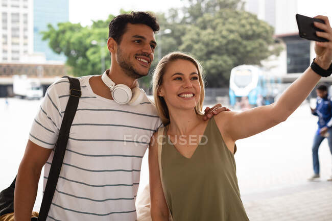 Vorderansicht eines glücklichen kaukasischen Paares, das tagsüber in den Straßen der Stadt unterwegs ist, auf der Straße steht und ein Selfie mit seinem Smartphone macht. — Stockfoto