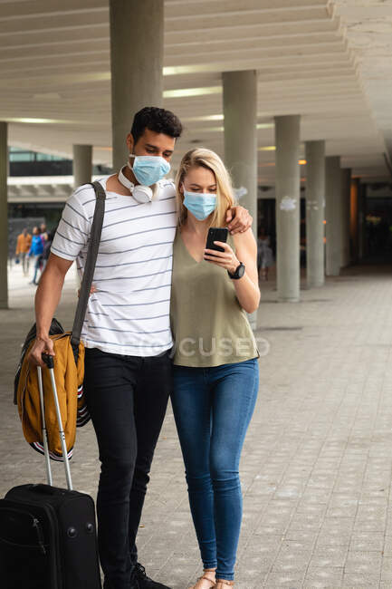 Vorderansicht eines kaukasischen Pärchens, das tagsüber auf den Straßen der Stadt unterwegs ist, Gesichtsmasken gegen Luftverschmutzung und Covid19 Coronavirus trägt und sein Smartphone benutzt.. — Stockfoto