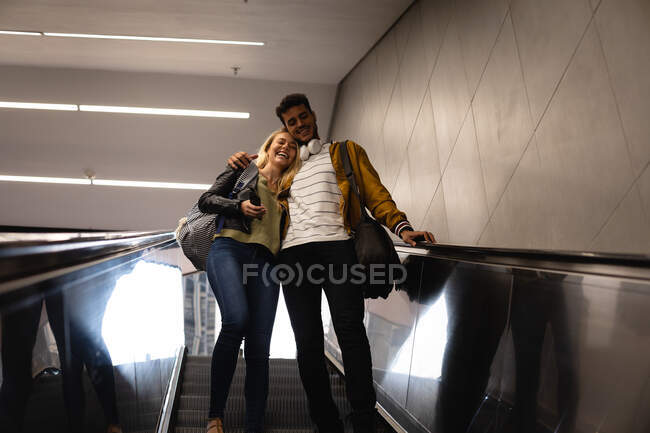 Vista frontal de ángulo bajo de una pareja caucásica en la ciudad, bajando en la estación de metro con una escalera mecánica, sonriendo y abrazando. - foto de stock