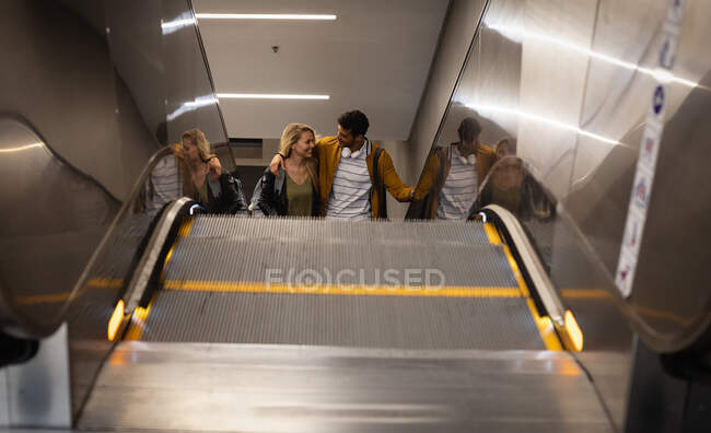 Vista frontal de ángulo alto de una pareja caucásica en la ciudad, subiendo en la estación de metro con una escalera mecánica, sonriendo y abrazando. - foto de stock