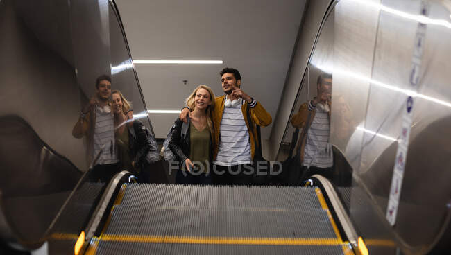 Vista frontal de ángulo bajo de una pareja caucásica en la ciudad, subiendo en la estación de metro con una escalera mecánica, sonriendo y abrazando. - foto de stock
