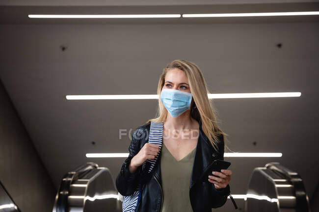 Vue de face d'une femme caucasienne aux longs cheveux aveugles, laissant un escalator, utilisant son smartphone et portant un masque facial contre la pollution atmosphérique et le coronavirus covid19. — Photo de stock