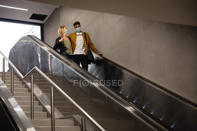 Vista frontal de baixo ângulo de um casal caucasiano na cidade, descendo na estação subterrânea com uma escada rolante, usando máscaras contra a poluição do ar e covid19 coronavírus . — Fotografia de Stock