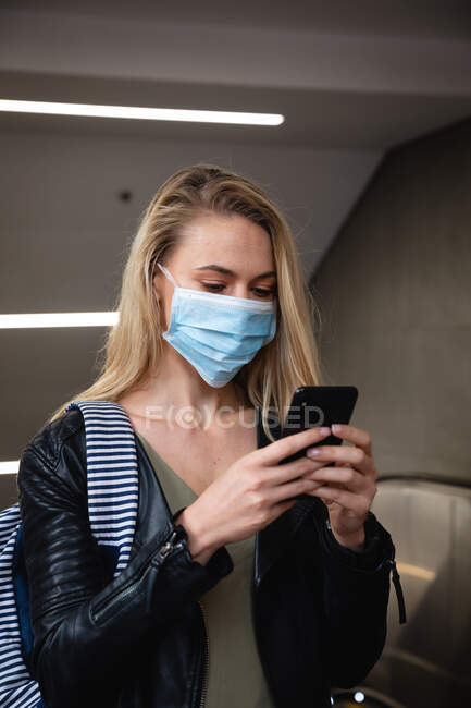 Вид спереди белокурой женщины с длинными светлыми волосами, покидающей эскалатор, использующей смартфон и маску для лица против загрязнения воздуха и вируса коронавируса covid19. — стоковое фото