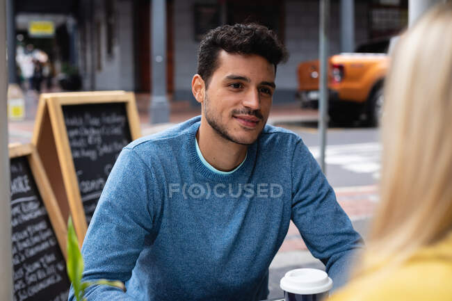 Vista frontal de un hombre caucásico de pelo corto y oscuro, sentado junto a una mesa en una terraza de café, disfrutando del tiempo con una mujer de pelo largo y rubio. - foto de stock