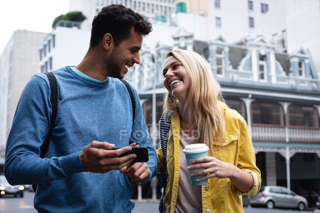 Vista frontale di una coppia caucasica felice in giro per le strade della città durante il giorno, con in mano una tazza di caffè da asporto, usando uno smartphone e sorridendo. — Foto stock
