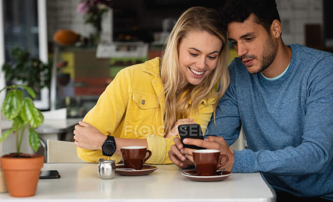 Vista frontal de una pareja caucásica sentada junto a una mesa en una cafetería, tomando café, disfrutando del tiempo juntos y usando su smartphone. - foto de stock