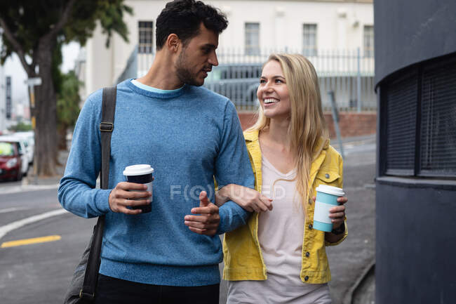 Vorderansicht eines glücklichen kaukasischen Paares, das in der Stadt unterwegs ist, Kaffee zum Mitnehmen in der Hand hält, Arm in Arm geht, lächelt und die gemeinsame Zeit genießt. — Stockfoto