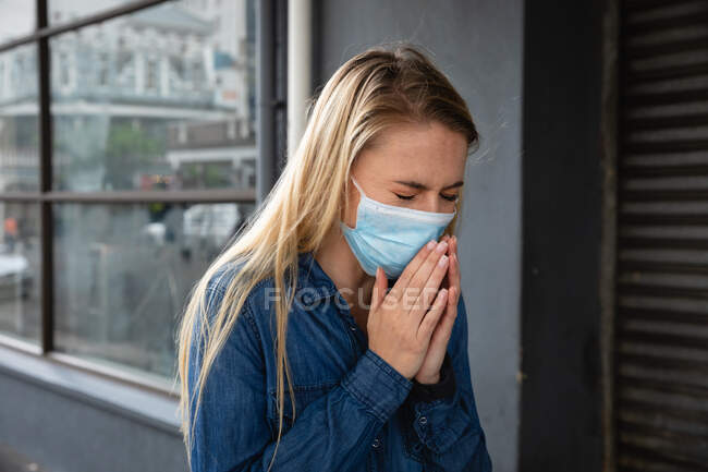 Vista frontal de cerca de una mujer caucásica con máscara facial contra la contaminación del aire y covid19 coronavirus, caminando por la calle y cubriéndose la cara mientras tose. - foto de stock