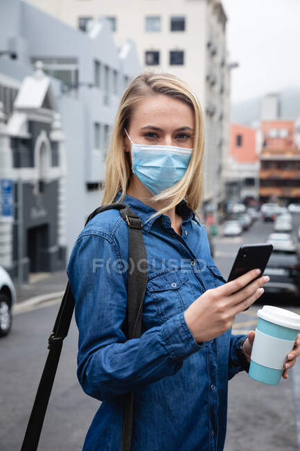 Vista frontale da vicino di una donna caucasica che indossa una maschera contro l'inquinamento atmosferico e covid19 coronavirus, passeggiando per le strade della città, usando il suo smartphone e tenendo in mano una tazza di caffè da asporto. — Foto stock