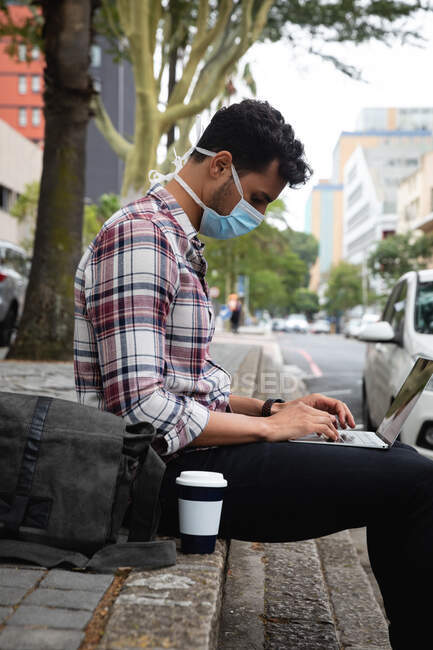Side view of a caucasian man wearing checkered shirt and face mask against air pollution and covid19 coronavirus, sitting in the stairs by the street and using his laptop computer. — Stock Photo