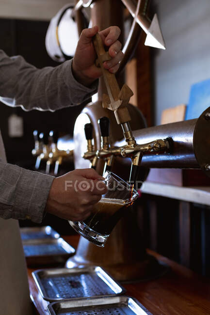 Partie médiane d'un homme travaillant dans un pub de microbrasserie, servant une pinte de bière, versant une boisson d'un robinet. — Photo de stock