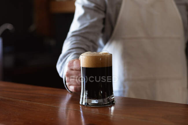 Mid section of man working at a microbrewery pub, wearing white apron, serving a pint of beer, putting it on a wooden bar. — Stock Photo