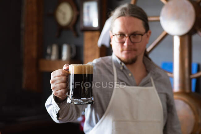 Homem caucasiano trabalhando em um pub de microcervejaria, vestindo avental branco, inspecionando uma caneca de cerveja, segurando-a na frente dele. — Fotografia de Stock