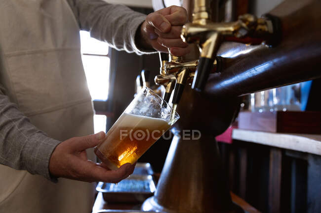 Parte média do homem que trabalha num pub de microcervejaria, servindo uma caneca de cerveja, derramando bebida de uma torneira. — Fotografia de Stock