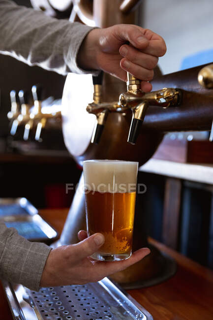 Mid section of man working at a microbrewery pub, serving a pint of beer, pouring beverage from a tap. — Stock Photo