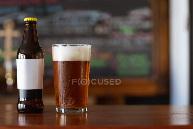 Una pinta di birra vera con una testa di schiuma e una bottiglia di vetro seduta sul bancone di legno in un pub di microbirreria. — Foto stock