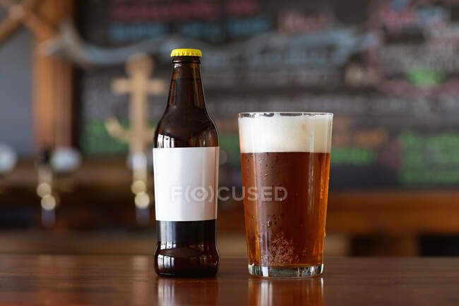 Una pinta di birra vera con una testa di schiuma e una bottiglia di vetro seduta sul bancone di legno in un pub di microbirreria. — Foto stock