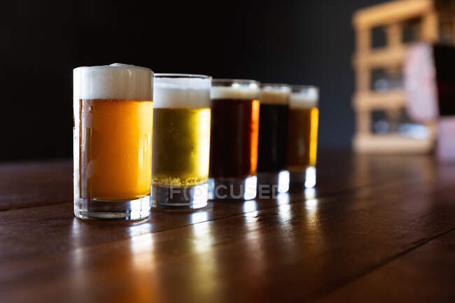 Five pints glasses of different kinds of beer with heads of foam sitting on the wooden bar at a microbrewery pub. — Stock Photo