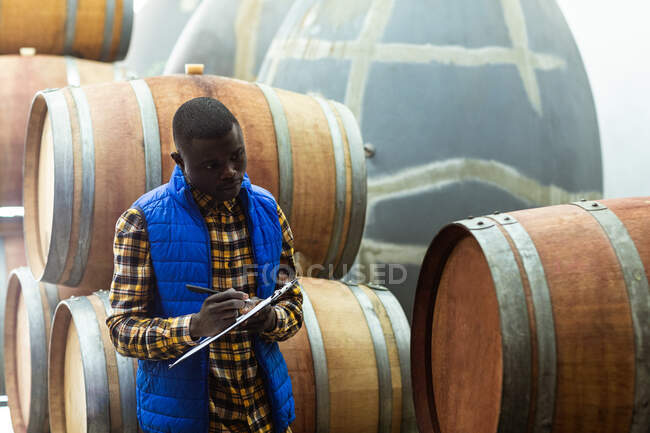 Uomo afroamericano che indossa un giubbotto blu, tiene un file e scrive numeri mentre conta i serbatoi in uno spazio di archiviazione in una microbirreria. — Foto stock