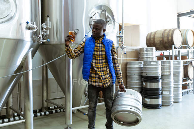 Hombre afroamericano trabajando en una microcervecería, sosteniendo una pinta de cerveza y un barril, con cubas y barriles de madera en el fondo. - foto de stock