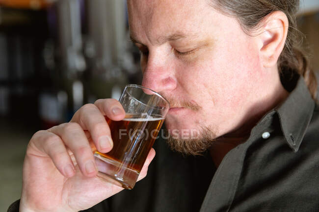 Primo piano di un uomo caucasico con i capelli lunghi che lavora in un microbirrificio, tiene un bicchiere di birra, lo annusa per l'ispezione. — Foto stock