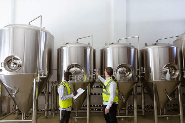 Caucasian and African American men working in a microbrewery, wearing high visibility vests, inspecting one of four vats set along the wall. — Stock Photo