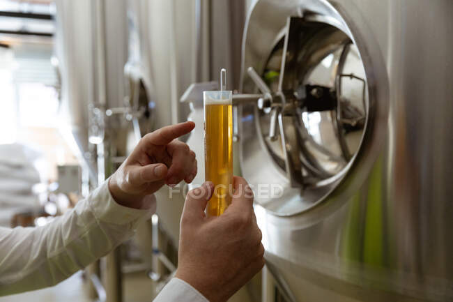 Средняя секция человека, работающего на микропивоварне, осматривающего стакан пива, проверяющего его цвет, с чанами на заднем плане. — стоковое фото