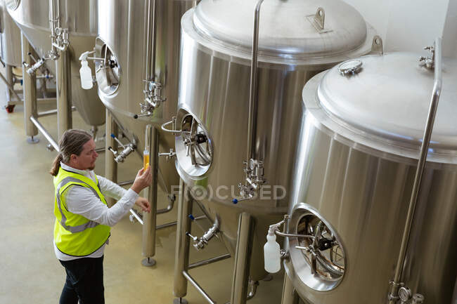 Hombre caucásico trabajando en una microcervecería, llevando un chaleco de alta visibilidad, inspeccionando un vaso de cerveza, comprobando su color. - foto de stock