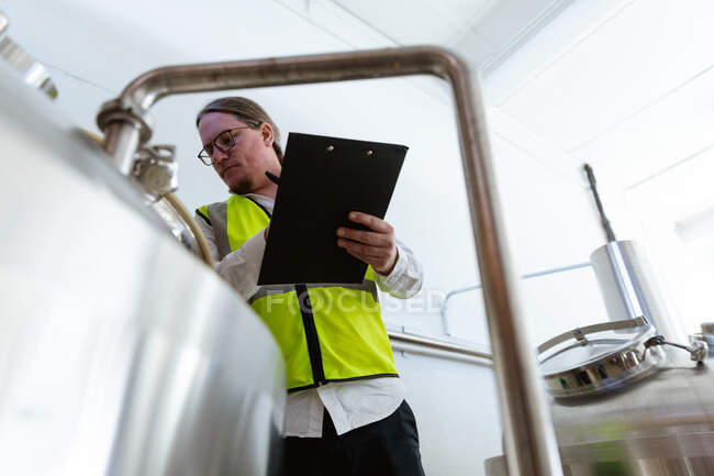 Low angle view of a Caucasian man wearing high visible vest, working in a microbrewery, holding a file and writing data while checking vats. — Stock Photo
