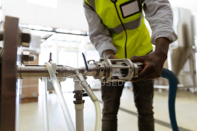 Vue du milieu de section d'un homme travaillant dans une microbrasserie, portant un gilet haute visibilité, inspectant un tuyau de réservoir. — Photo de stock