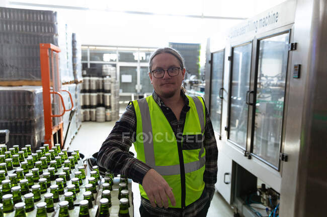 Портрет кавказького чоловіка, який носить високу видимість жилета, працює в мікропивоварні, спираючись на пляшки, готові до доставки і дивлячись прямо в камеру.. — стокове фото