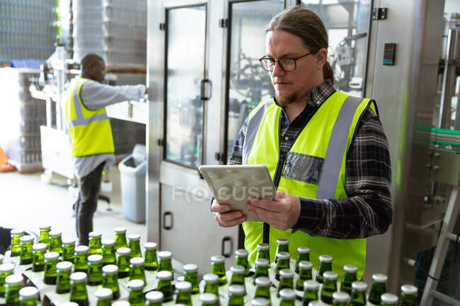 Caucasian man wearing high visibility vest, working in a microbrewery, using a tablet while checking bottles of beer prepared for delivery with an African American man working in the background. — Stock Photo