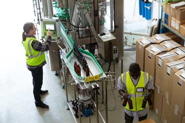 High angle view of a Caucasian and African American men wearing high visibility vest, working in a microbrewery, checking bottles of beer. — Stock Photo