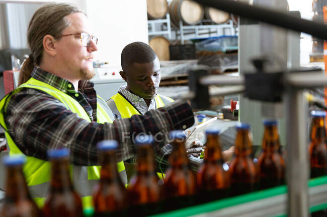 Кавказький чоловік у високій видимості, працює у мікропивоварні, перевіряє пляшки пива з афроамериканцем, який працює на задньому плані.. — стокове фото