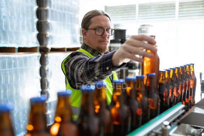 Homme caucasien vêtu d'un gilet de haute visibilité, travaillant dans une microbrasserie, vérifiant les bouteilles de bière en verre foncé sur un tapis roulant. — Photo de stock