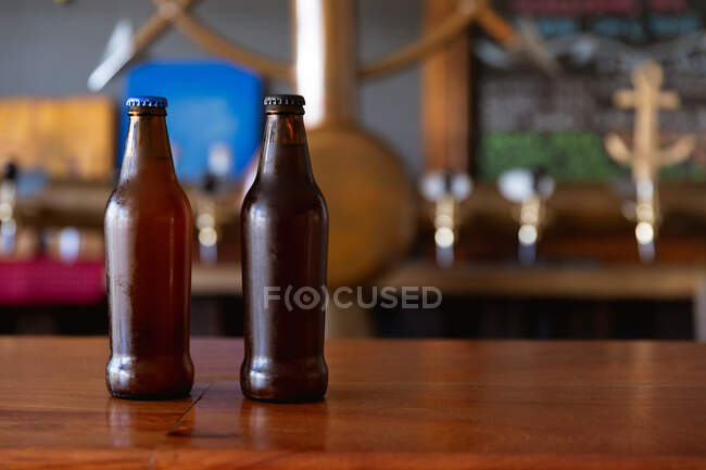 Zwei dunkle Glasflaschen mit Bierdeckeln sitzen auf der hölzernen Theke einer Mikrobrauerei mit Zapfhähnen im Hintergrund. — Stockfoto
