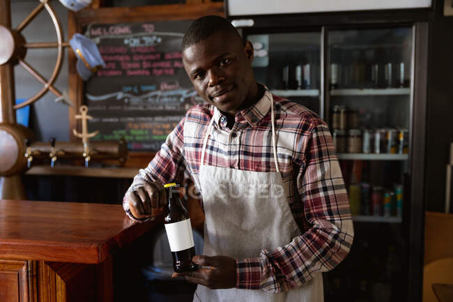 Hombre afroamericano trabajando en un pub de microcervecería, usando delantal blanco, sosteniendo una botella de cerveza y mirando directamente a una cámara. - foto de stock