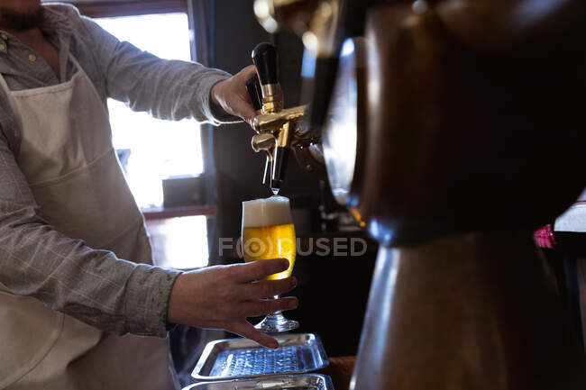 Parte média do homem que trabalha num pub de microcervejaria, servindo uma caneca de cerveja, derramando bebida de uma torneira. — Fotografia de Stock