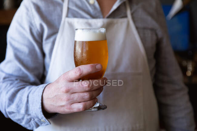 Partie médiane d'un homme travaillant dans un pub de microbrasserie, portant un tablier blanc, servant une pinte de bière, la tenant devant lui. — Photo de stock