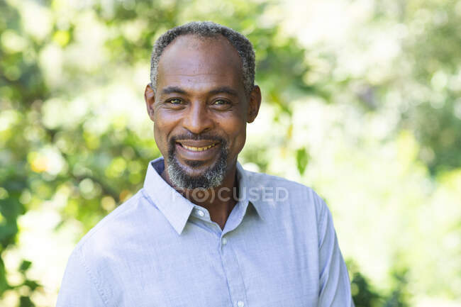 Portrait d'un bel homme afro-américain âgé profitant de sa retraite, dans un jardin au soleil regardant la caméra sourire, s'isolant lui-même lors d'une pandémie de coronavirus covid19 — Photo de stock