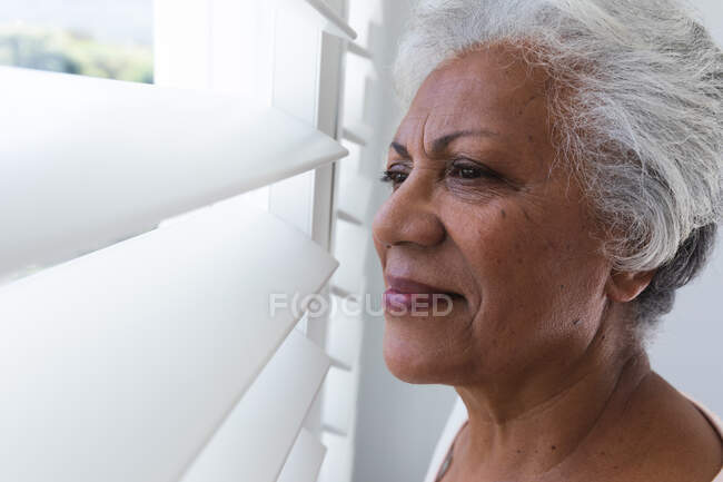 Вигляд збоку поруч приваблива пенсіонерка афро-американська жінка з коротким білим волоссям вдома дивлячись у вікно між білими гучнішими віконцями в сонячний літній день і посміхаючись — стокове фото