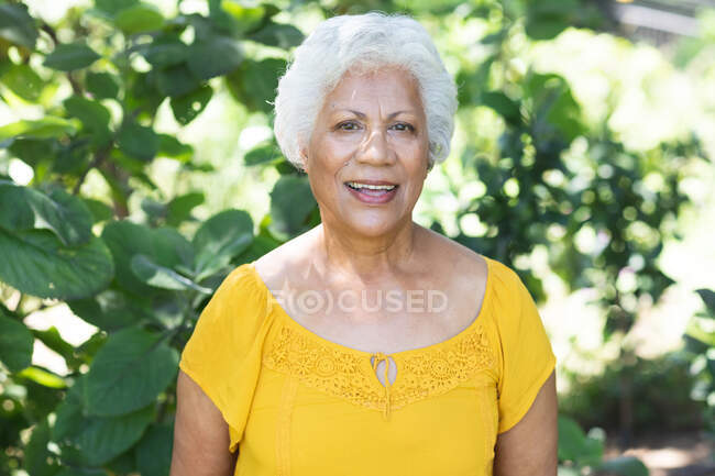 Retrato de una atractiva mujer afroamericana mayor con el pelo blanco corto disfrutando de su retiro en un jardín al sol, mirando a la cámara y sonriendo, auto aislándose durante la pandemia de coronavirus covid19 - foto de stock