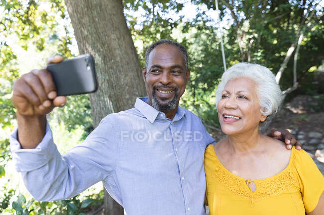 Um casal afro-americano sênior desfrutando de sua aposentadoria, de pé em um jardim ao sol abraçando e sorrindo, o homem segurando um smartphone e tirando uma selfie — Fotografia de Stock