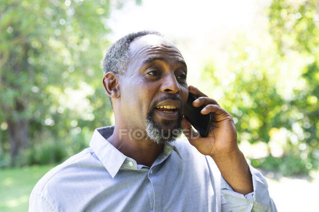 Ein gut aussehender älterer afroamerikanischer Mann genießt seinen Ruhestand, in einem Garten in der Sonne, telefoniert mit einem Handy und lächelt — Stockfoto